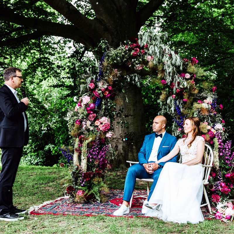 Trauredner und Hochzeitspaar sitzt vor bunt geschmücktem Baum